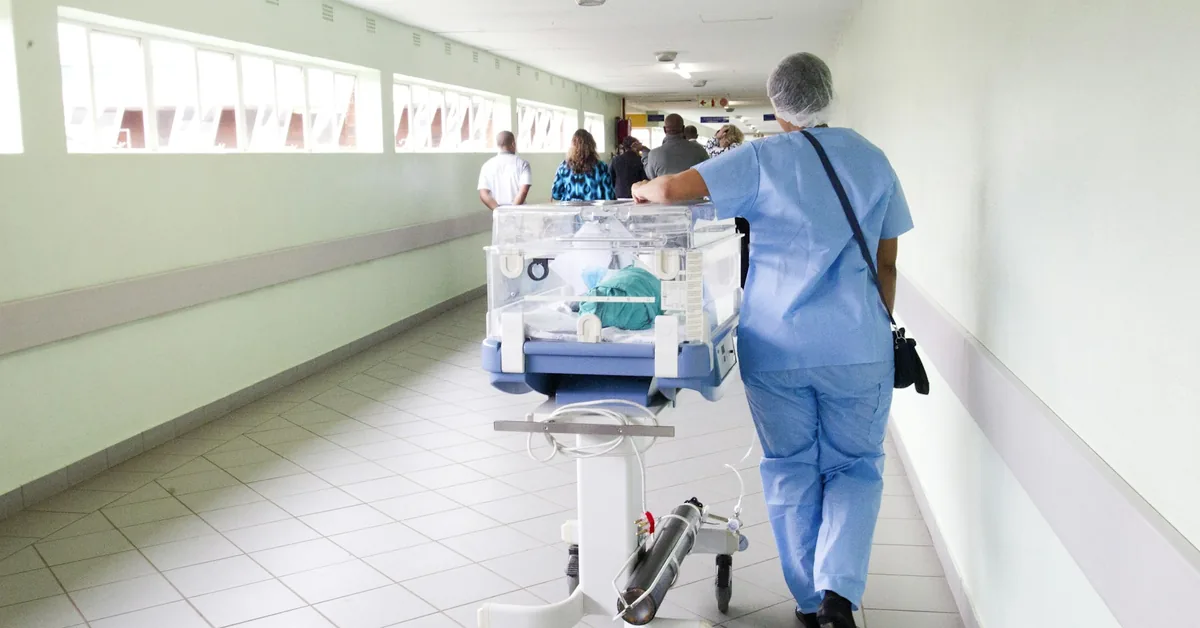 A nurse in scrubs walking a long tiled hospital corridor, pushing an incubator trolley
